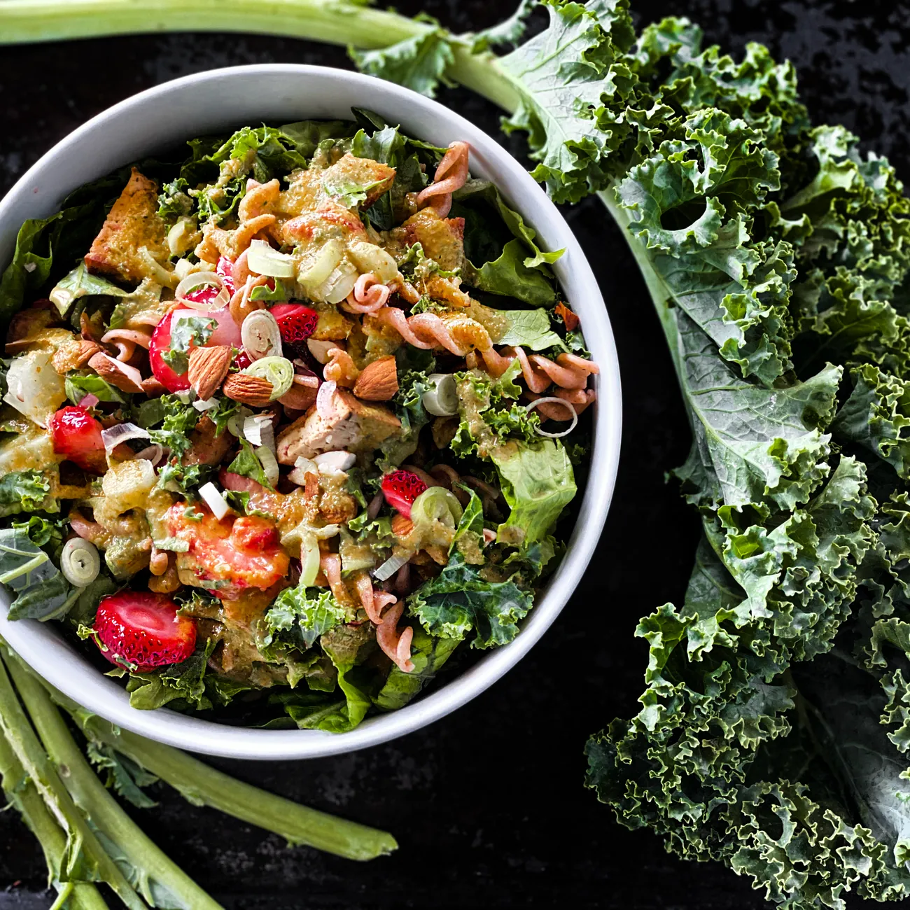 Bát salad cải xoăn với cà chua và hạt, quanh là lá cải trên nền tối